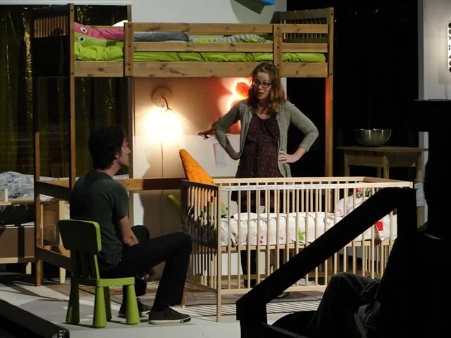 Two people, a woman and a man, are in a room with a wooden bunk bed and a crib. The woman is standing and talking to the man, who is sitting on a green chair. The room is decorated with warm lighting, and there is a lamp on the wall.